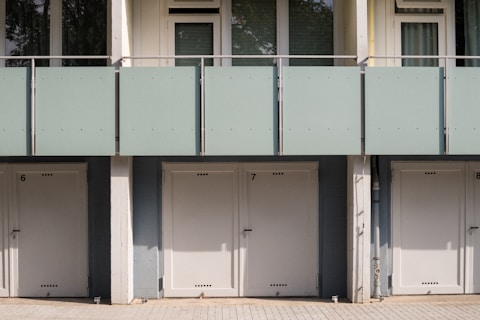 A series of closed garage doors numbered 6, 7, and 8 are positioned side by side beneath a modern apartment building. Above the doors, there is a glass balcony with a light green frosted panel railing and windows with shades partially drawn. The setting is likely urban and has a clean, minimalistic architectural design.