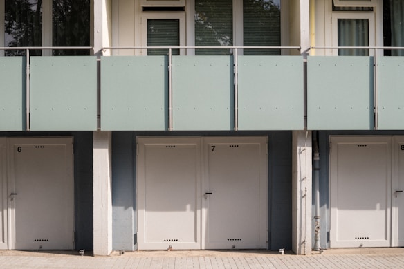 A series of closed garage doors numbered 6, 7, and 8 are positioned side by side beneath a modern apartment building. Above the doors, there is a glass balcony with a light green frosted panel railing and windows with shades partially drawn. The setting is likely urban and has a clean, minimalistic architectural design.