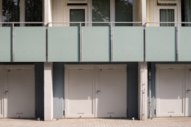A series of closed garage doors numbered 6, 7, and 8 are positioned side by side beneath a modern apartment building. Above the doors, there is a glass balcony with a light green frosted panel railing and windows with shades partially drawn. The setting is likely urban and has a clean, minimalistic architectural design.