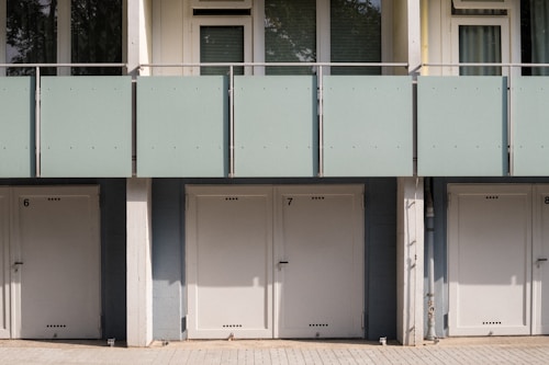 A series of closed garage doors numbered 6, 7, and 8 are positioned side by side beneath a modern apartment building. Above the doors, there is a glass balcony with a light green frosted panel railing and windows with shades partially drawn. The setting is likely urban and has a clean, minimalistic architectural design.