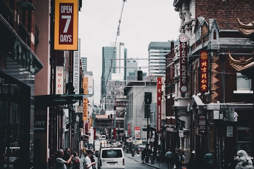 A bustling city street features various shops with vibrant signage, including restaurants and businesses offering goods. The architecture blends modern high-rises in the background with older buildings in the foreground, some adorned with decorative elements. Pedestrians are walking alongside parked cars, contributing to the lively urban atmosphere.