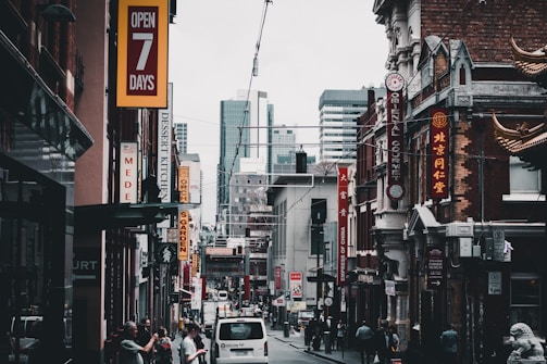 A bustling city street features various shops with vibrant signage, including restaurants and businesses offering goods. The architecture blends modern high-rises in the background with older buildings in the foreground, some adorned with decorative elements. Pedestrians are walking alongside parked cars, contributing to the lively urban atmosphere.