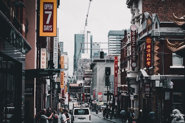 A bustling city street features various shops with vibrant signage, including restaurants and businesses offering goods. The architecture blends modern high-rises in the background with older buildings in the foreground, some adorned with decorative elements. Pedestrians are walking alongside parked cars, contributing to the lively urban atmosphere.
