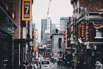 A bustling city street features various shops with vibrant signage, including restaurants and businesses offering goods. The architecture blends modern high-rises in the background with older buildings in the foreground, some adorned with decorative elements. Pedestrians are walking alongside parked cars, contributing to the lively urban atmosphere.