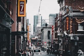 A bustling city street features various shops with vibrant signage, including restaurants and businesses offering goods. The architecture blends modern high-rises in the background with older buildings in the foreground, some adorned with decorative elements. Pedestrians are walking alongside parked cars, contributing to the lively urban atmosphere.