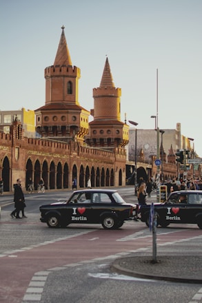 A historic brick bridge with two prominent towers, each topped with a conical roof, spans across a busy street. Numerous pedestrians and cyclists can be seen moving along the pathways. Two classic cars, each with an 'I ♥ Berlin' decal, are driving on the street below. The late afternoon sun casts warm tones across the scene, giving the architecture a golden glow.