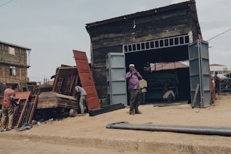 A rugged workshop setting with men working on metal structures outside and inside a large wooden shed. One man stands outside talking on the phone, while others are busy with metal doors and frames. The area is surrounded by dirt and debris, with a few buildings visible in the background.