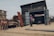 A rugged workshop setting with men working on metal structures outside and inside a large wooden shed. One man stands outside talking on the phone, while others are busy with metal doors and frames. The area is surrounded by dirt and debris, with a few buildings visible in the background.