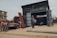 A rugged workshop setting with men working on metal structures outside and inside a large wooden shed. One man stands outside talking on the phone, while others are busy with metal doors and frames. The area is surrounded by dirt and debris, with a few buildings visible in the background.