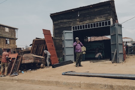 A rugged workshop setting with men working on metal structures outside and inside a large wooden shed. One man stands outside talking on the phone, while others are busy with metal doors and frames. The area is surrounded by dirt and debris, with a few buildings visible in the background.