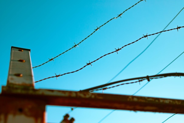 Barbed wire fencing set against a clear blue sky with a metal pole on the left side. The wires are rusted, giving an aged and weathered appearance.