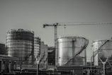 A series of large industrial storage tanks with a crane positioned in the background. Each tank is metal with curved staircases wrapping around the exterior. The scene looks industrial and is in a monochromatic color scheme.
