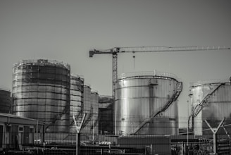 A series of large industrial storage tanks with a crane positioned in the background. Each tank is metal with curved staircases wrapping around the exterior. The scene looks industrial and is in a monochromatic color scheme.