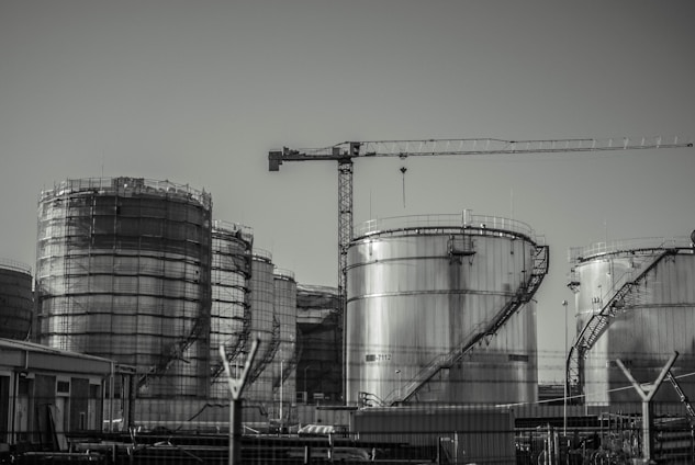 A series of large industrial storage tanks with a crane positioned in the background. Each tank is metal with curved staircases wrapping around the exterior. The scene looks industrial and is in a monochromatic color scheme.
