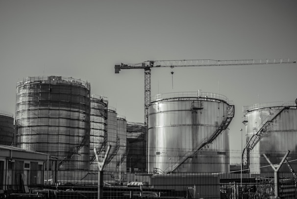 A series of large industrial storage tanks with a crane positioned in the background. Each tank is metal with curved staircases wrapping around the exterior. The scene looks industrial and is in a monochromatic color scheme.