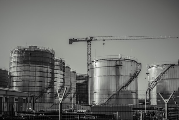 A series of large industrial storage tanks with a crane positioned in the background. Each tank is metal with curved staircases wrapping around the exterior. The scene looks industrial and is in a monochromatic color scheme.