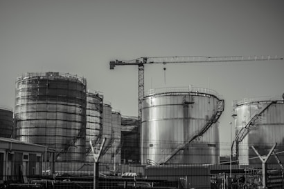 A series of large industrial storage tanks with a crane positioned in the background. Each tank is metal with curved staircases wrapping around the exterior. The scene looks industrial and is in a monochromatic color scheme.