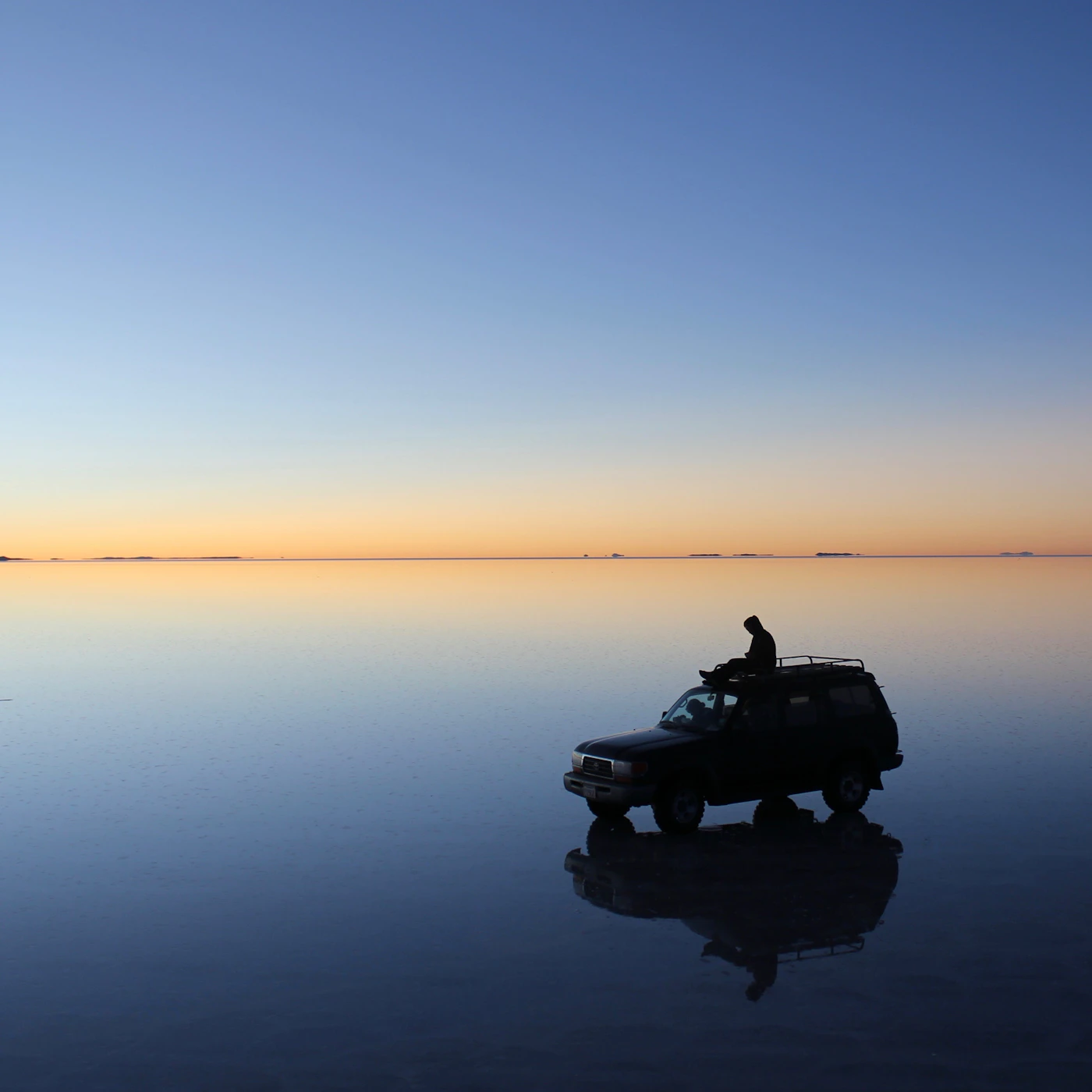 A lone vehicle silhouetted at sunset on the mirror-like surface of the Salar de Uyuni salt flat