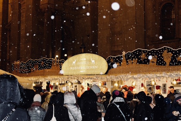 A bustling outdoor market stall with a sign reading 'Winter Wonder Kitchen' adorned with festive decorations and lights. People are gathered, wearing winter clothing, which indicates cold weather. Snowflakes are visible in the air, enhancing the wintery atmosphere. The background features a large, illuminated building.