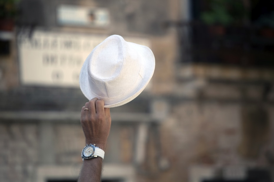 A hand is holding up a white hat against a blurred urban background. The person is wearing a wristwatch and a ring.