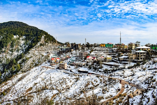 A small town nestled in a hilly, snow-covered landscape. Colorful houses are scattered across the slopes, with a winding road leading through the area. Dense forested hills rise in the background under a clear blue sky with scattered clouds.