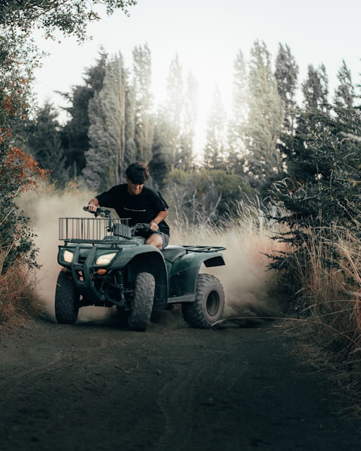 A person is riding a green all-terrain vehicle (ATV) on a dirt path surrounded by tall grass and trees. Dust is being kicked up as the ATV moves, indicating motion and speed. The background shows a dense forest with tall trees and a somewhat bright, cloudy sky.