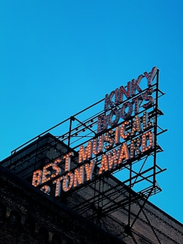 A large metal sign mounted on a brick building displays text with bulb lighting. The words 'Kinky Boots', 'Best Musical', and '6 Tony Award' are prominently featured. The sky in the background is a clear blue.