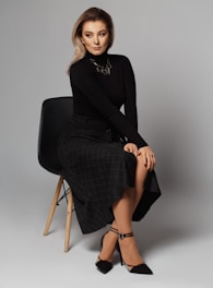 A woman with long hair and poised demeanor sits on a modern wooden chair. She wears a black turtleneck sweater, a dark checkered skirt, and black high-heeled shoes. A geometric necklace accessorizes her outfit. Her hands rest gracefully on her lap, and her expression is one of calm confidence.