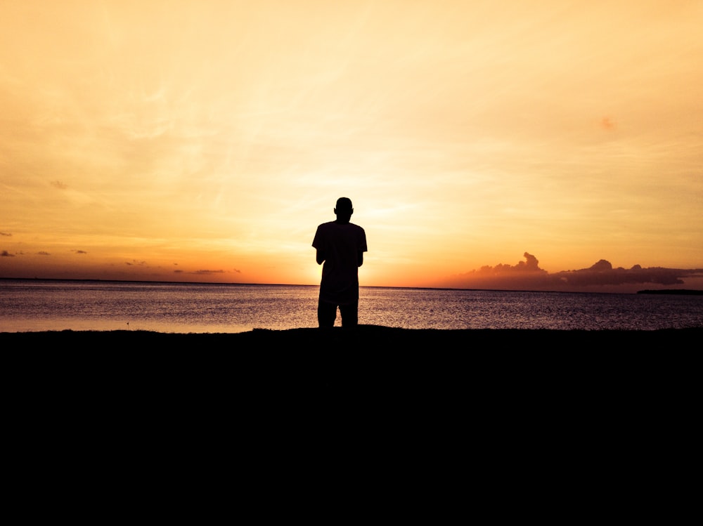 Foto da silhueta da pessoa em pé na frente do corpo de água durante o pôr do sol