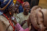 A group of people dressed in traditional African attire gather together, with a man at the center smiling broadly. The clothing features vibrant patterns, and some women wear ornate head wraps. Ornamental beads and accessories are visible, suggesting a festive or ceremonial gathering.