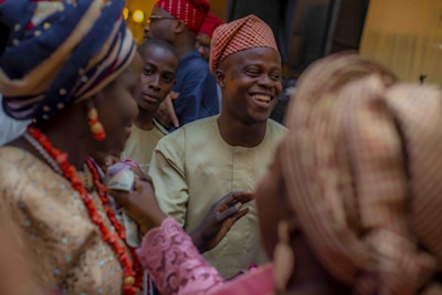 A group of people dressed in traditional African attire gather together, with a man at the center smiling broadly. The clothing features vibrant patterns, and some women wear ornate head wraps. Ornamental beads and accessories are visible, suggesting a festive or ceremonial gathering.