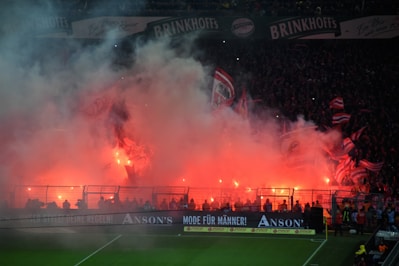 A large crowd of people in a stadium is surrounded by dense smoke and illuminated by bright red flares. The scene is lively with numerous flags waving, and the smoke adds a dramatic and intense atmosphere. The stadium banners and fencing are visible along with the illuminated stands filled with spectators.