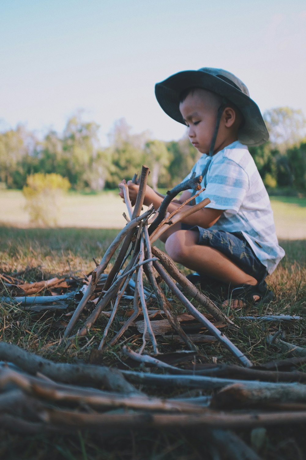 boy squat sitting beside wooden sticks formed to a pyramid during day