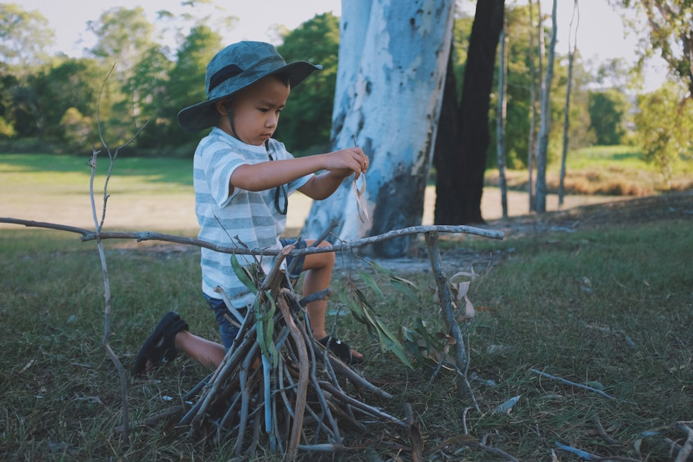 boy wearing gray hat, white-and-gray striped crew-neck t-shirt and gray shorts kneeling on ground playing twigs during daytime