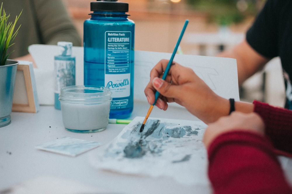 person holding blue paint brush painting on paper on table