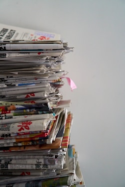 A tall stack of newspapers piled neatly against a plain background, with some publications appearing to show colorful images and text in a foreign language.