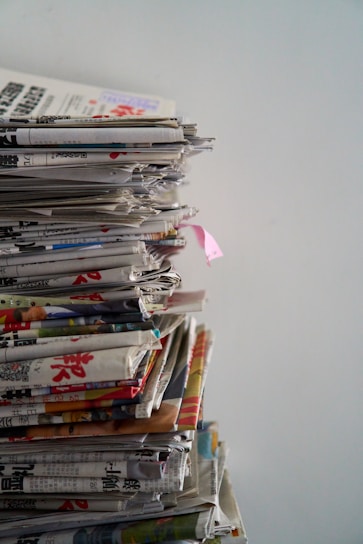 A tall stack of newspapers piled neatly against a plain background, with some publications appearing to show colorful images and text in a foreign language.