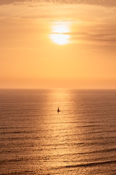 A serene seascape at sunset with a vibrant golden sky and reflections on the calm ocean waters. A small sailboat is visible in the distance, silhouetted against the bright backdrop.