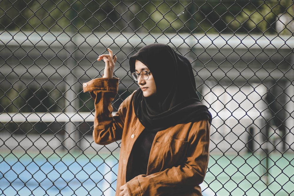 selective focus photography of woman wearing black hijab scarf and brown coat leaning on metal chain-link fence during daytime