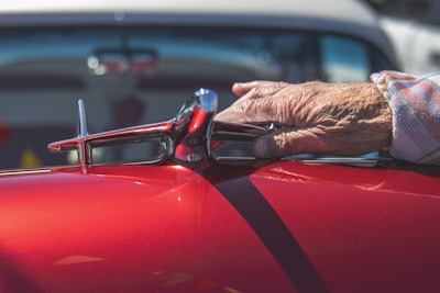An elderly hand rests on a shiny red classic car hood, near a chrome hood ornament shaped like an airplane, with a softly focused background featuring a glimpse of another vehicle.