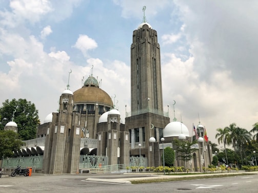 A grand architectural structure featuring a combination of domes and a tall tower, adorned with Islamic crescents. The building is surrounded by lush green trees and a clear sky with some clouds. Flags and a few motorcycles are visible near the entrance, and there are manicured shrubs lining the pathway.