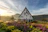 A picturesque landscape featuring a quaint A-frame house with a large triangular roof, surrounded by lush greenery and vibrant flowers in the foreground. The background features a serene lake, a rolling hill, and a forest under a partly cloudy sky during what appears to be sunrise or sunset.
