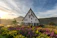 A picturesque landscape featuring a quaint A-frame house with a large triangular roof, surrounded by lush greenery and vibrant flowers in the foreground. The background features a serene lake, a rolling hill, and a forest under a partly cloudy sky during what appears to be sunrise or sunset.