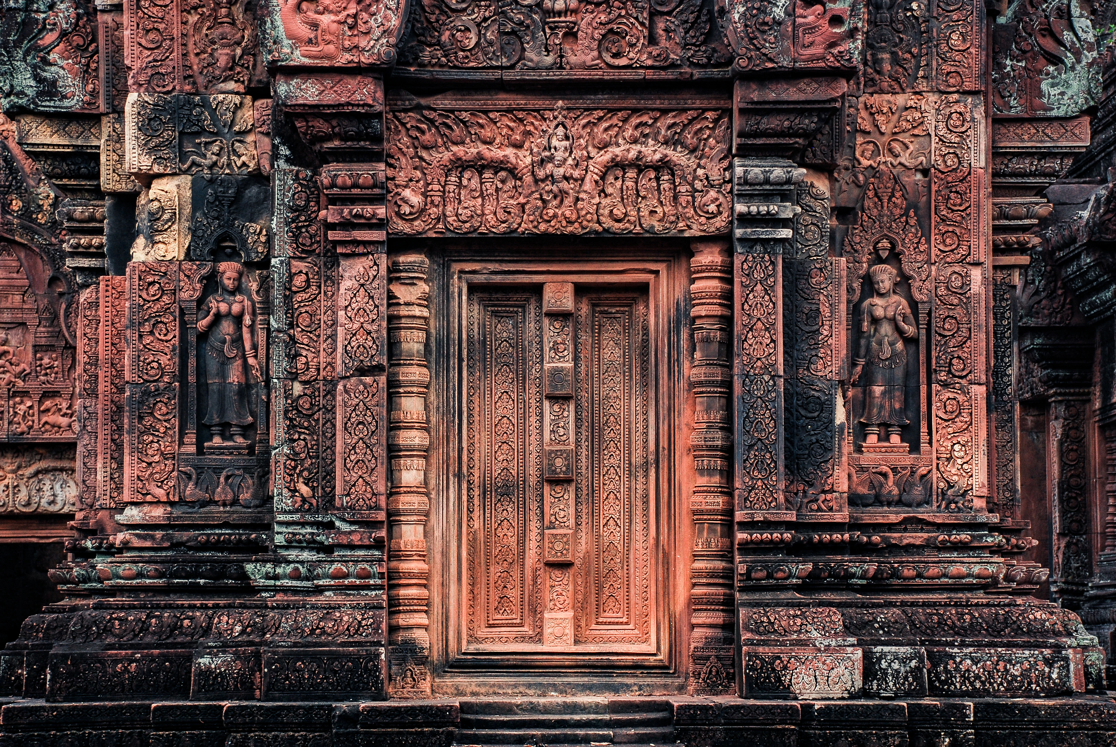 The pink sandstone false door of Banteay Srei, flanked by two carved guardian devatas standing in their niches beneath a densely carved pediment of foliage and figures