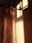 Warm sunlight casts shadows through a window onto a tiled wall next to a hanging curtain. The curtain features a subtle geometric pattern, with soft folds highlighted by the light.