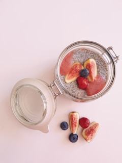 A mason jar filled with a chia seed pudding and topped with fresh figs, strawberries, and blueberries. The jar is open and placed on a light surface. Beside it, some fruit pieces are arranged, including whole blueberries and halved figs.