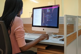 A person with long hair is seated at a desk, working on a computer. The computer displays lines of code, suggesting programming or software development activity. The workspace includes a modern cubicle setup with wooden and glass elements.