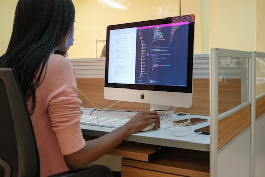 A person with long hair is seated at a desk, working on a computer. The computer displays lines of code, suggesting programming or software development activity. The workspace includes a modern cubicle setup with wooden and glass elements.