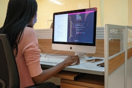 A person with long hair is seated at a desk, working on a computer. The computer displays lines of code, suggesting programming or software development activity. The workspace includes a modern cubicle setup with wooden and glass elements.