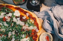 A freshly baked pizza topped with arugula, slices of parmesan cheese, and tomato sauce is placed on a wooden surface. Beside the pizza, there is a dark cloth napkin, a small bowl of coarse salt with a red spoon, and a can of olive oil. The pizza has a crispy crust and vibrant toppings that give it an appetizing look.